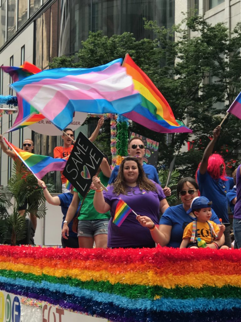 Image depicting at a Pride event, holding the lgbtq+ rainbow flag and the trans flag.